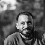 A candid black and white portrait of a smiling man with a beard, set against a blurred outdoor background.